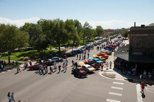 Aerial shot of vintage cars parked on a quaint main street between shops and a tree-filled park. People are milling about.